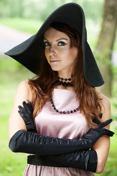 Beautiful young woman in a big black hat walks in the park, vintage style, elegance.