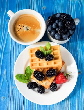 İyi sabahlar. Böğürtlenli ev yapımı waffle, çilek ve kahvaltıda bir fincan kahve mavi ahşap arka planda..