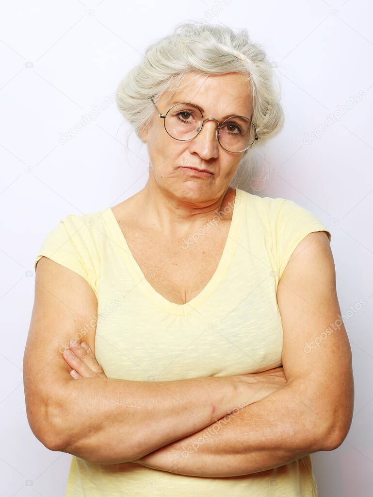 The disgruntled grandmother looks up and condemns. An elderly woman stands with her arms crossed over her chest. Shot over grey background.