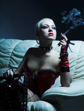 Göz kamaştırıcı hanımefendi, kanepede oturup sigara içerken kölesini bekliyor. Fetiş ve yetişkin oyunları.