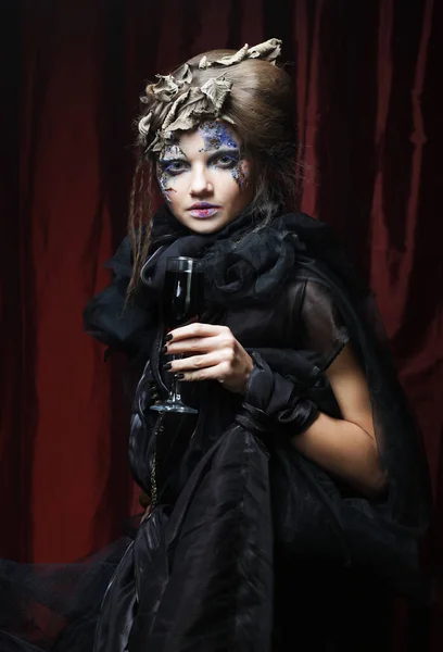 Party, holiday and Halloween concept. A beautiful woman in a witch costume with bright makeup and curly hair is holding a glass of wine.