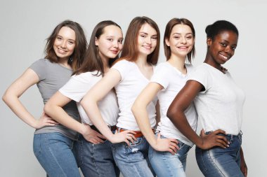 Farklı boylarda ve etnik kökenlere sahip bir grup mutlu genç kadın gri arkaplanda tişört giyiyor.