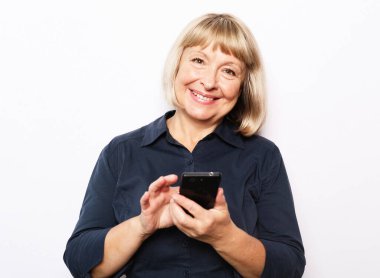 Tehnology and old people concept: Pretty senior female in casual outfit using modern smartphone standing on white background. Old woman watching funny videos on internet.