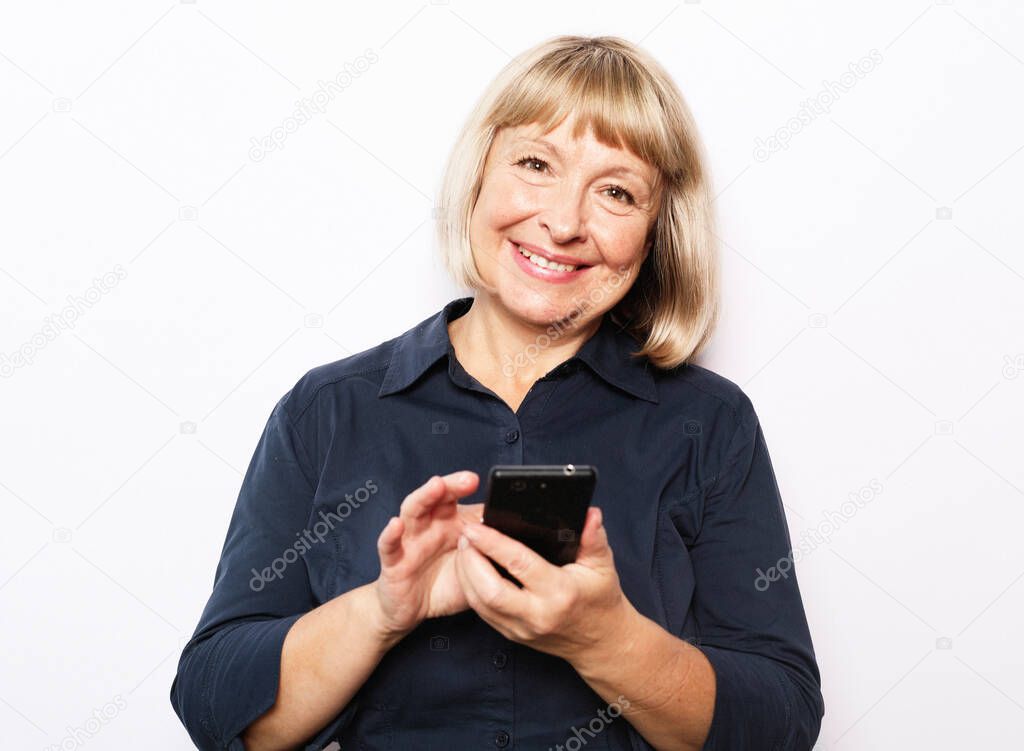Tehnology and old people concept: Pretty senior female in casual outfit using modern smartphone standing on white background. Old woman watching funny videos on internet.