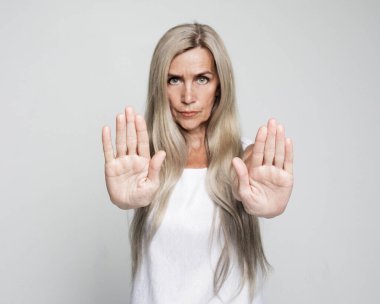 Yaşlı gri saçlı kadın, gri arka planda ciddi ve kendinden emin bir ifadeyle dur işareti yapıyor.