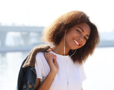 Genç Afrikalı Amerikalı kız cep telefonu ve kulaklık kullanıyor, gülümsüyor ve denize yakın duruyor.