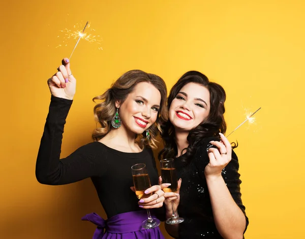 Sarı arka planda şampanya bardakları ve maytapları olan iki genç kadının portresi. — Stok fotoğraf