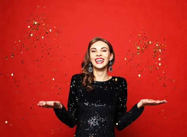 Концепция праздников и вечеринок: красивая девушка в вечернем платье, стоящая под дождём конфетти на красном фоне — стоковое фото