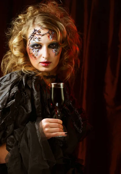 Piękna kobieta w stroju czarownicy z jasnym makijażem i kręconymi włosami trzyma kieliszek wina. — Zdjęcie stockowe