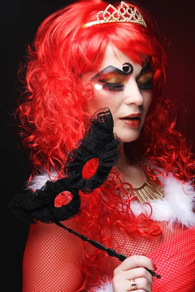 Encantadora mujer juguetona con peluca rizada roja sostiene una máscara y sonríe. Carnaval, fiesta y celebración. —  Fotos de Stock
