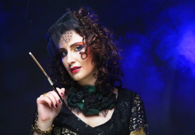 Parlak makyajlı, duvak takmış, elinde sigara tutan güzel bir kadın.