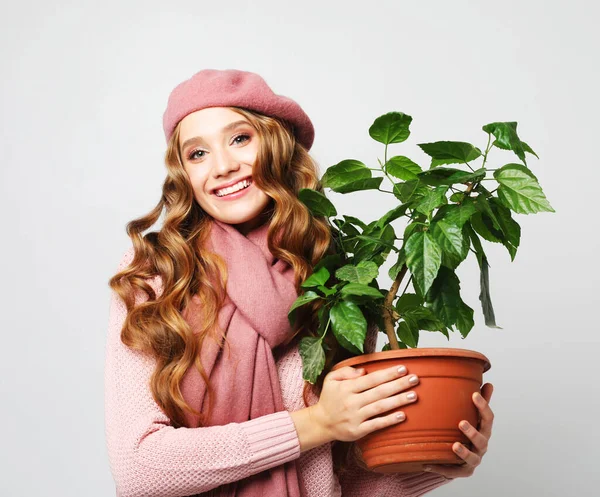 Wanita tersenyum dengan rambut panjang bergelombang mengenakan pakaian merah muda, berdiri di atas latar belakang abu-abu dan memegang bunga dalam pot — Stok Foto