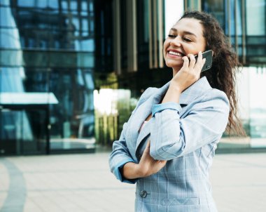 İş merkezinin arka planında iş tarzı giyinmiş genç bir Afrikalı kadın elinde bir cep telefonu tutuyor.