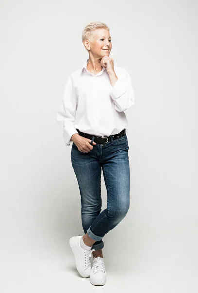 Full längd foto av elegant äldre kvinna bär jeans och vit blus med kort vitt hår poserar på en ljusgrå bakgrund. — Stockfoto