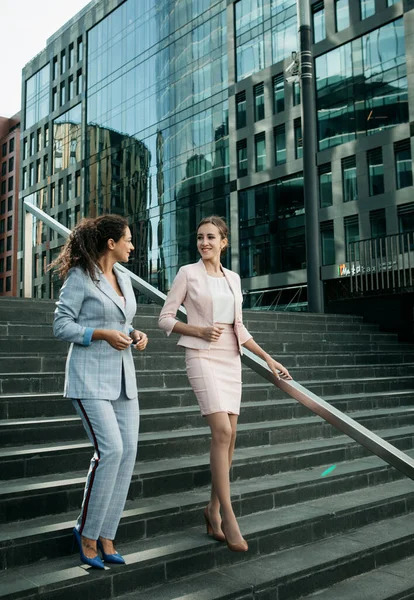 Two smiling business women talking while walking down stairs near modern office building. — Stock Photo, Image