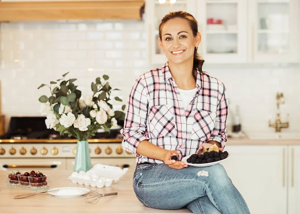 A fiatal, gyönyörű, boldog nő ül a konyhaasztalnál, és egy tál sütit tart a kezében.. — Stock Fotó