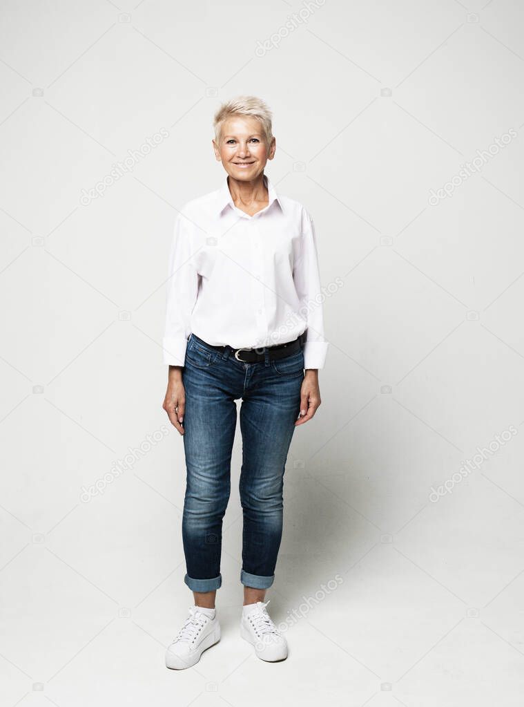 Full length photo of stylish elderly woman wearing jeans and white blouse with short white hair posing on a light gray background.
