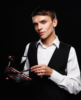 Genç erkek makyaj sanatçısı elinde gölge paleti ve fırça tutuyor..