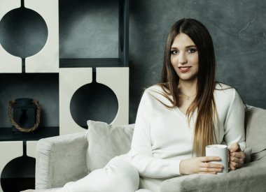 Güzel ve sevimli. Elinde kahve fincanı tutan ve evdeki sandalyeye oturan güzel genç bir kadın. Yaşam tarzı kavramı.