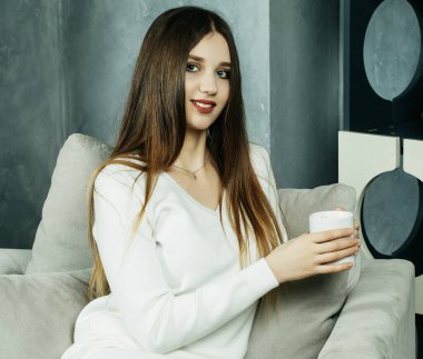 Güzel ve sevimli. Elinde kahve fincanı tutan ve evdeki sandalyeye oturan güzel genç bir kadın. Yaşam tarzı kavramı.