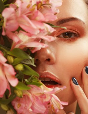 Güzel, genç bir kadın. Makyajı yumuşak renklerde. Kocaman pembe bir çiçek tutuyor.