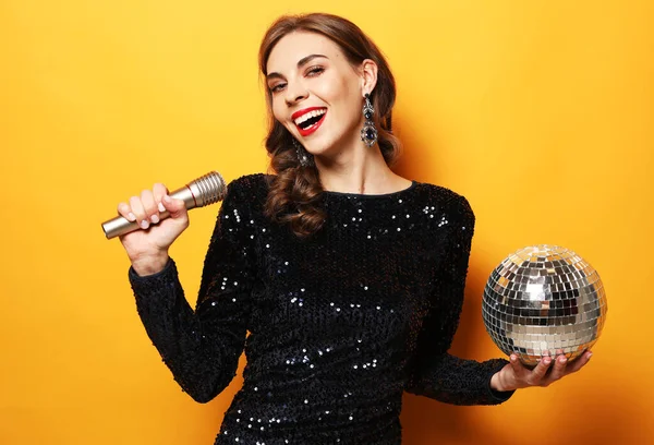 Young brunette woman with long curly hair dressed in evening dress holding a microphone and disco ball — Stock Photo, Image