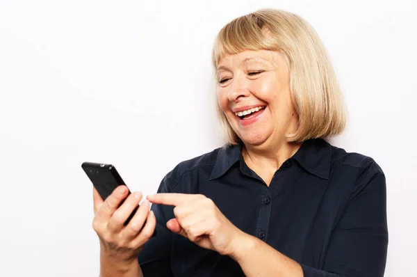 穿着休闲装的漂亮老年女性使用现代智能手机，站在白色背景下大声大笑。老太婆在网上看滑稽录像. — 图库照片