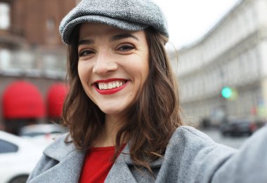 Gri şapkalı, ceketli, güzel bir kadın eski binanın önünden geçerken selfie çekiyor..