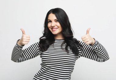 Vücut dili, duygular ve insanlar konsepti: Çizgili kazaklı genç esmer kadın gülümsüyor ve başparmağını kaldırıyor