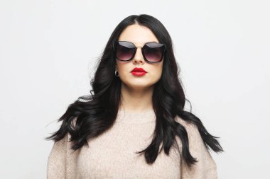 Pembe kazak ve güneş gözlüğü takan genç bir kadın gri arka planda poz veriyor.