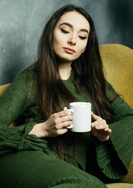 Güzel ve sevimli. Elinde kahve fincanı tutan ve evdeki sandalyeye oturan güzel genç bir kadın. Yaşam tarzı ve insan kavramı.