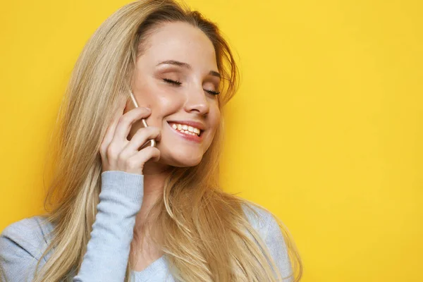 年轻的金发女人穿着蓝色衬衫，在黄色背景下通过电话交谈 — 图库照片