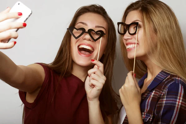 Duas jovens mulheres felizes com óculos de festa tirando selfie com celular sobre fundo branco — Fotografia de Stock