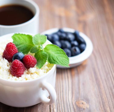 Beyaz kasede bir fincan kahve ve süzme peynir ahududu ve ahşap arka planda yaban mersini. Sağlıklı Kahvaltı. Diyet için iyi..