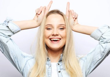 Beyaz arka plan üzerine gündelik giyinmiş genç, güzel sarışın kadın boynuz gibi parmağıyla komik jestler yapıyor.