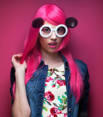 Pembe saçlı, pembe arka planda büyük güneş gözlüklü, modaya uygun genç bir kadın..