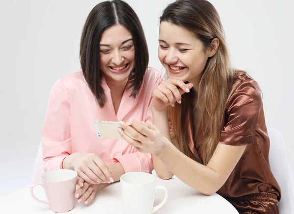 Vackra flickvänner i pyjamas sitter vid bordet, dricker te eller kaffe och pratar i ett socialt nätverk. — Stockfoto