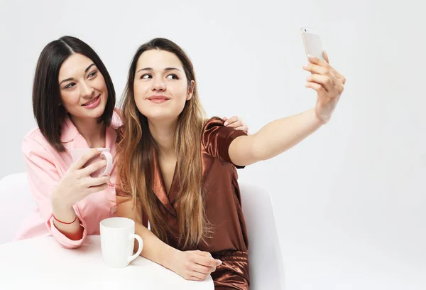 Девушки со смартфоном делают селфи и пьют кофе на белом фоне — стоковое фото