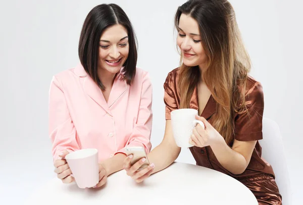 Красиві подружки в піжамі сидять за столом, п'ють чай або каву і спілкуються в соціальній мережі . — стокове фото