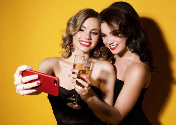 Duas jovens mulheres chiques em vestidos de cocktail preto beber champanhe e tirar uma selfie. — Fotografia de Stock
