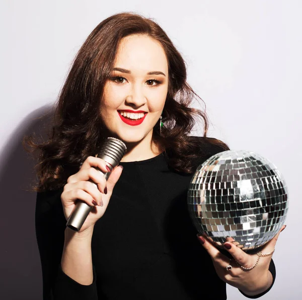 Young brunette woman wearing black dress, holding disco ball and singing into microphone — Stock Photo, Image