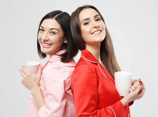 Duas belas jovens amigas vestidas de pijama vermelho e rosa segurando uma xícara com café — Fotografia de Stock