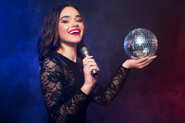 Lifestyle, party and people concept: young woman wearing black dress, holding disco ball and singing into microphone over dark background — Stock Photo, Image
