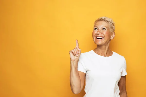 Senior grey-haired woman wearing white t-shirt pointing finger up with successful idea over yellow background. — Stock Photo, Image