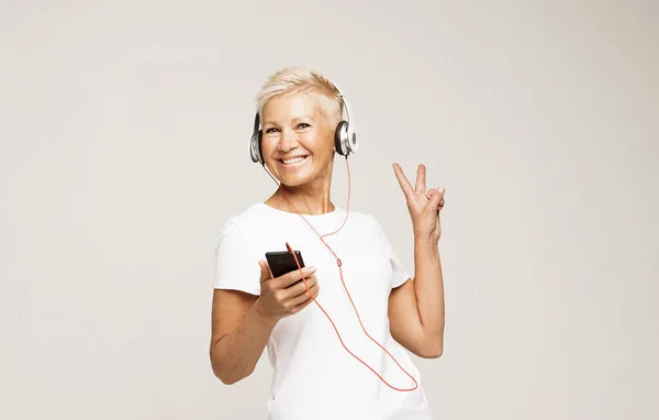 Donna con i capelli bianchi corti ascoltare musica con le cuffie e mostrare segno di vittoria su sfondo grigio — Foto Stock