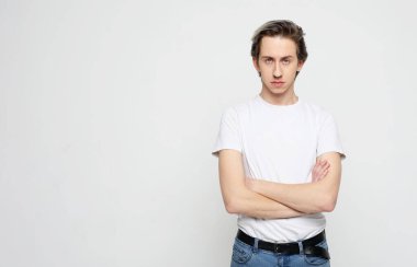 Beyaz tişörtlü genç adam el ele tutuşuyor ve gri arka planda kameraya bakıyor.