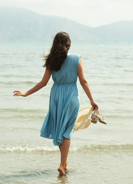 青いドレスを着た若いブルネットの女性の背中の景色は ビーチを裸足で歩き 水の中で足をぶら下げています ハッピーバケーション — ストック写真