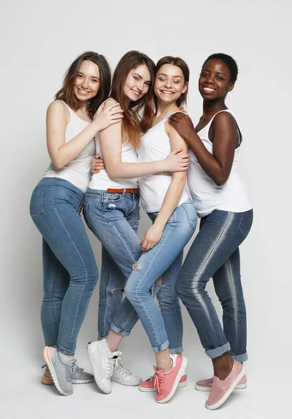 Retrato de cuatro hermosas mujeres sonrientes en jeans de pie juntas aisladas sobre un fondo gris, abrazándose. —  Fotos de Stock