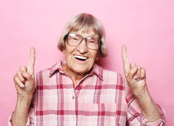 Retrato de feliz sorrindo mulher idosa usando óculos apontando o dedo para cima sobre fundo rosa — Fotografia de Stock
