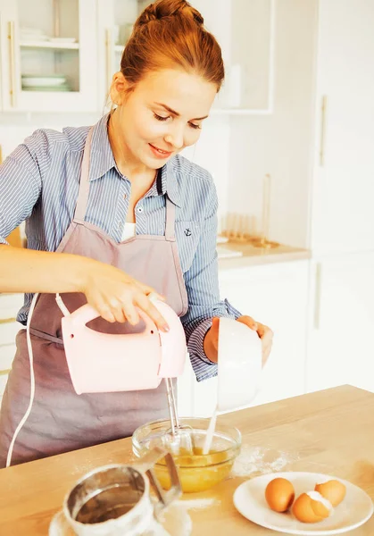 Young woman baking a cake in the kitchen, using a mixer to whisk the fresh ingredients in a glass mixing bowl. — Stock Photo, Image